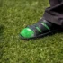 Close-up of a green socked foot in a black sandal resting on grass, with a dark pant leg nearby.