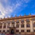 Historic stone building with ornate statues along the roofline beneath a bright blue sky with wispy clouds.