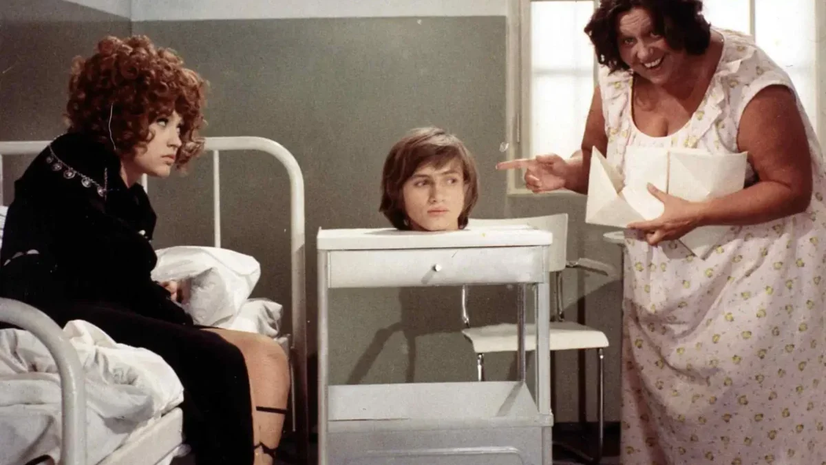 Nurse in a floral gown gestures toward a head in a display container on a wheeled cart, while a patient sits up in bed watching.