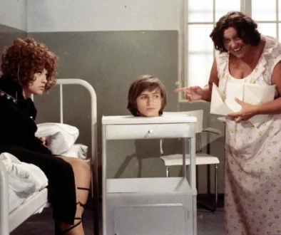 Nurse in a floral gown gestures toward a head in a display container on a wheeled cart, while a patient sits up in bed watching.