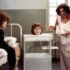 Nurse in a floral gown gestures toward a head in a display container on a wheeled cart, while a patient sits up in bed watching.