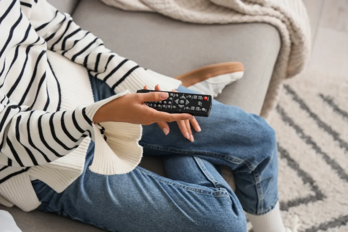 Žena sedí na gauči a v ruce drží TV ovladač.