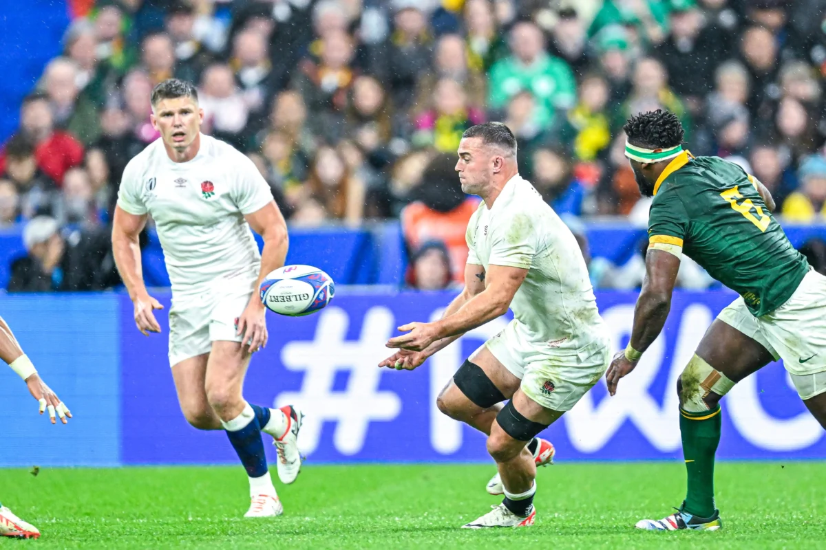 Rugby players in white and green jerseys chase a ball on a grassy field with a blurred crowd in the stands behind them.