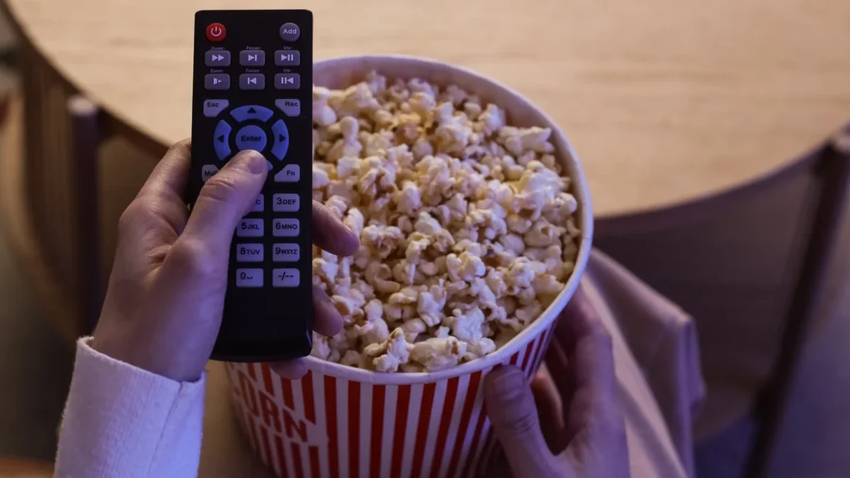 Hand holding a TV remote over a large bucket of popcorn, as if about to press a button to start the movie.