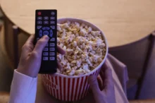 Hand holding a TV remote over a large bucket of popcorn, as if about to press a button to start the movie.