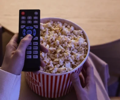 Hand holding a TV remote over a large bucket of popcorn, as if about to press a button to start the movie.