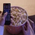 Hand holding a TV remote over a large bucket of popcorn, as if about to press a button to start the movie.