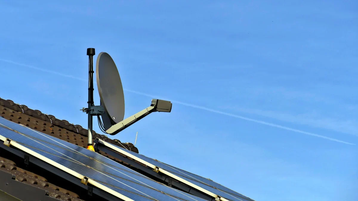 Satellite dish mounted on a pitched roof beside solar panels against a clear blue sky.