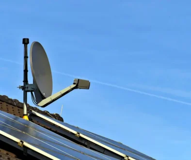 Satellite dish mounted on a pitched roof beside solar panels against a clear blue sky.