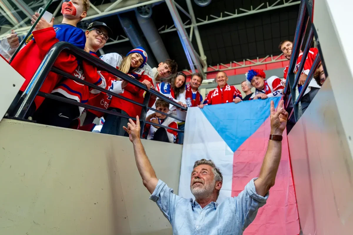 Older man with a gray beard raises his arms toward a cheering group on a balcony, a Czech flag behind them? visible flag colors blue, white, red.