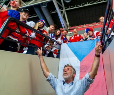 Older man with a gray beard raises his arms toward a cheering group on a balcony, a Czech flag behind them? visible flag colors blue, white, red.