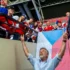 Older man with a gray beard raises his arms toward a cheering group on a balcony, a Czech flag behind them? visible flag colors blue, white, red.