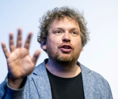 Man with light brown hair and beard speaks while raising an open hand toward the camera.