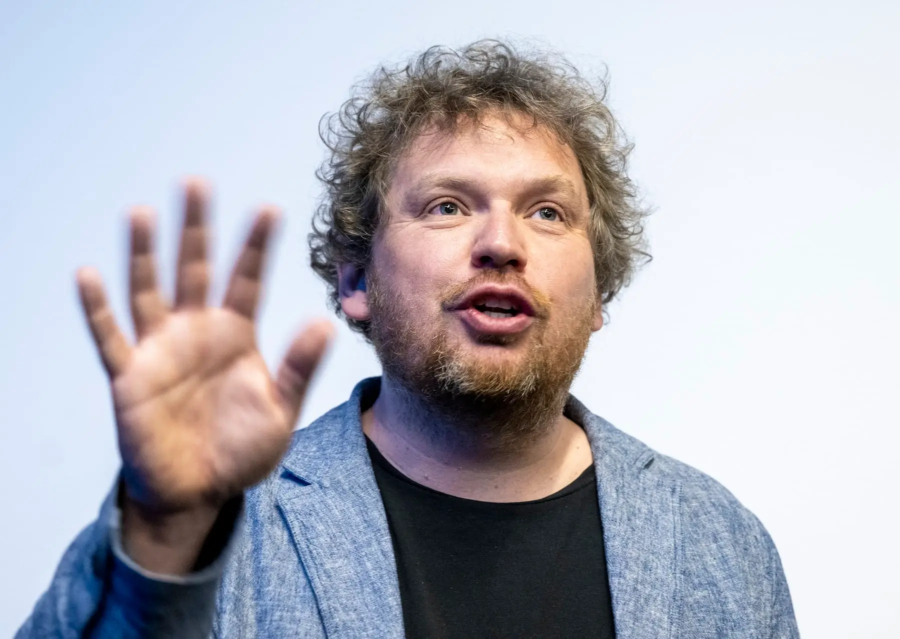 Man with light brown hair and beard speaks while raising an open hand toward the camera.