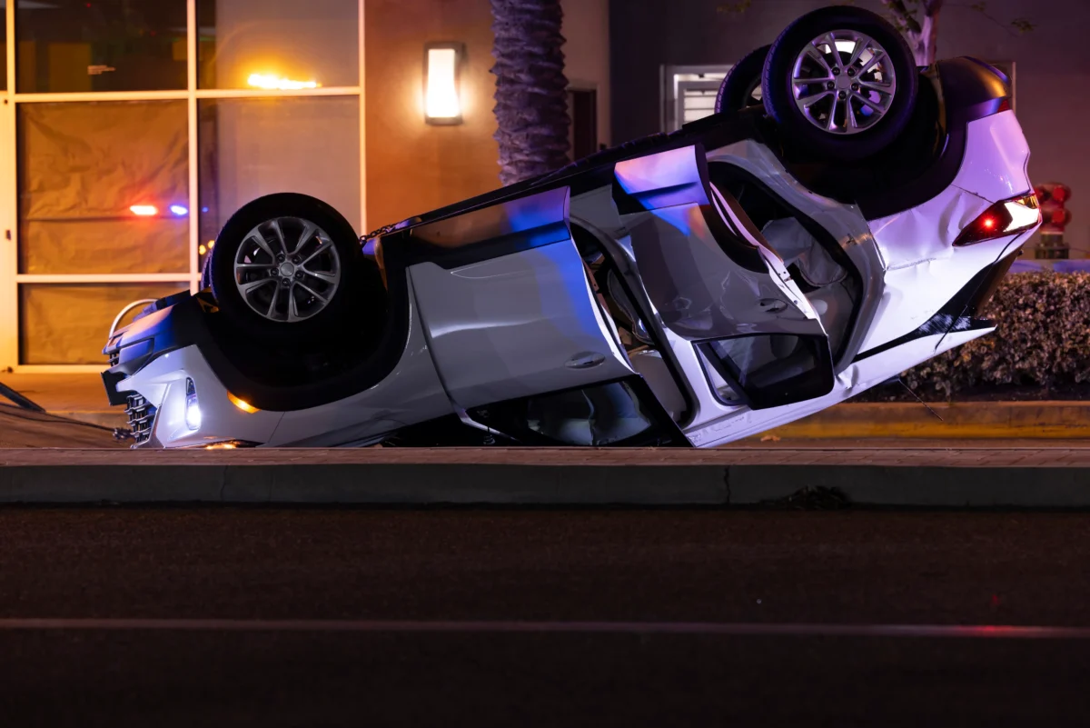 White SUV upside down on a street at night, with visible damage and wheels in the air near a lit building exterior.
