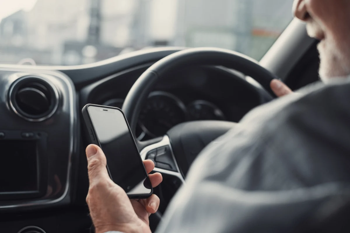 Person driving a car while holding a smartphone in their hand, steering wheel in view.