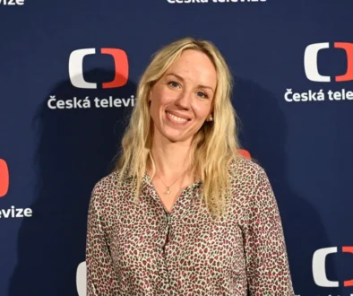 Petra Hřebíčková stojí před nápisy Česká televize a usmívá se.