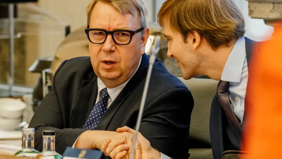 Two businessmen in suits sit at a conference table, one speaking toward the camera while the other leans in to listen.