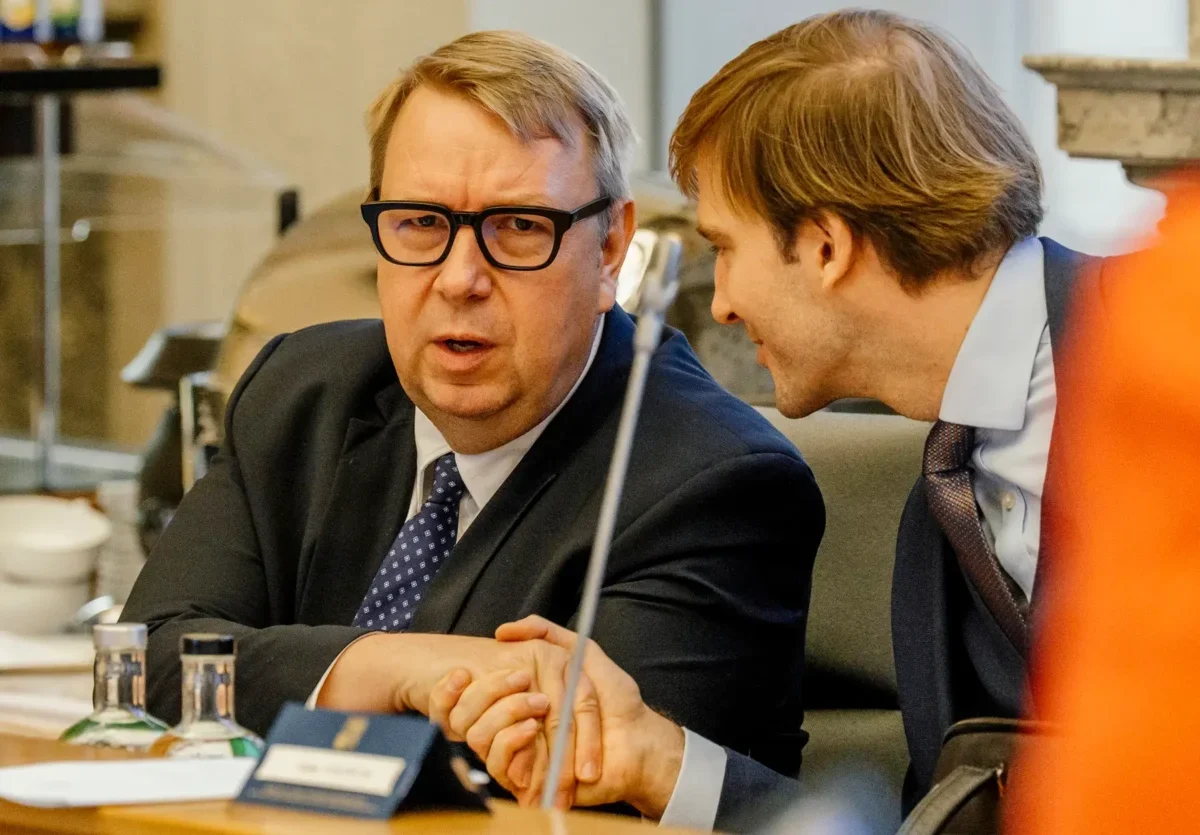 Two businessmen in suits sit at a conference table, one speaking toward the camera while the other leans in to listen.