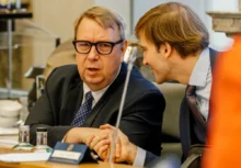 Two businessmen in suits sit at a conference table, one speaking toward the camera while the other leans in to listen.