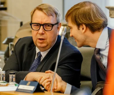 Two businessmen in suits sit at a conference table, one speaking toward the camera while the other leans in to listen.