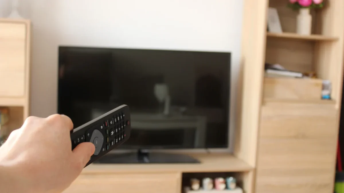 Person’s hand holds a TV remote, aimed at a flat-screen TV in a wood-furnished living room start foreground.