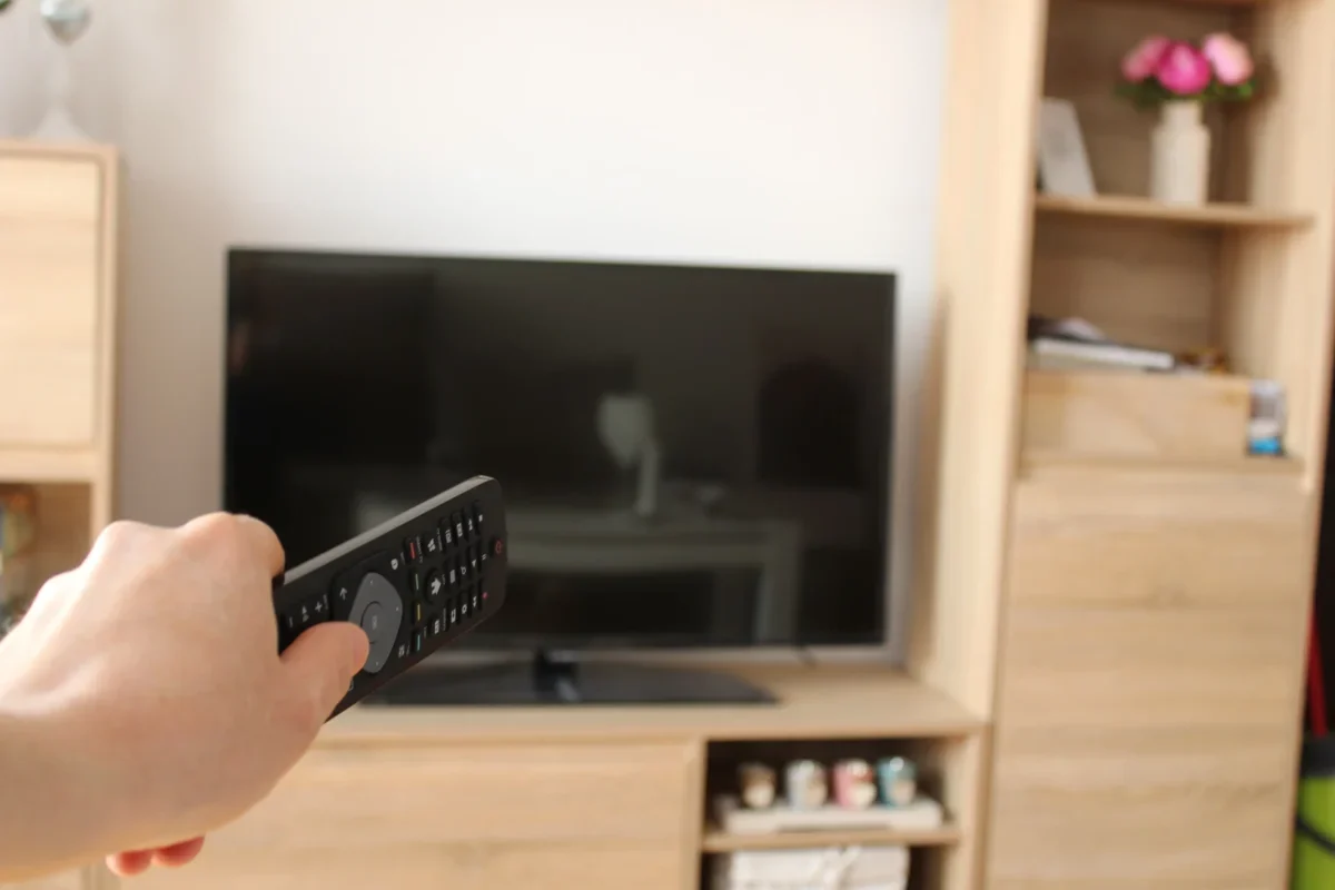 Person’s hand holds a TV remote, aimed at a flat-screen TV in a wood-furnished living room start foreground.