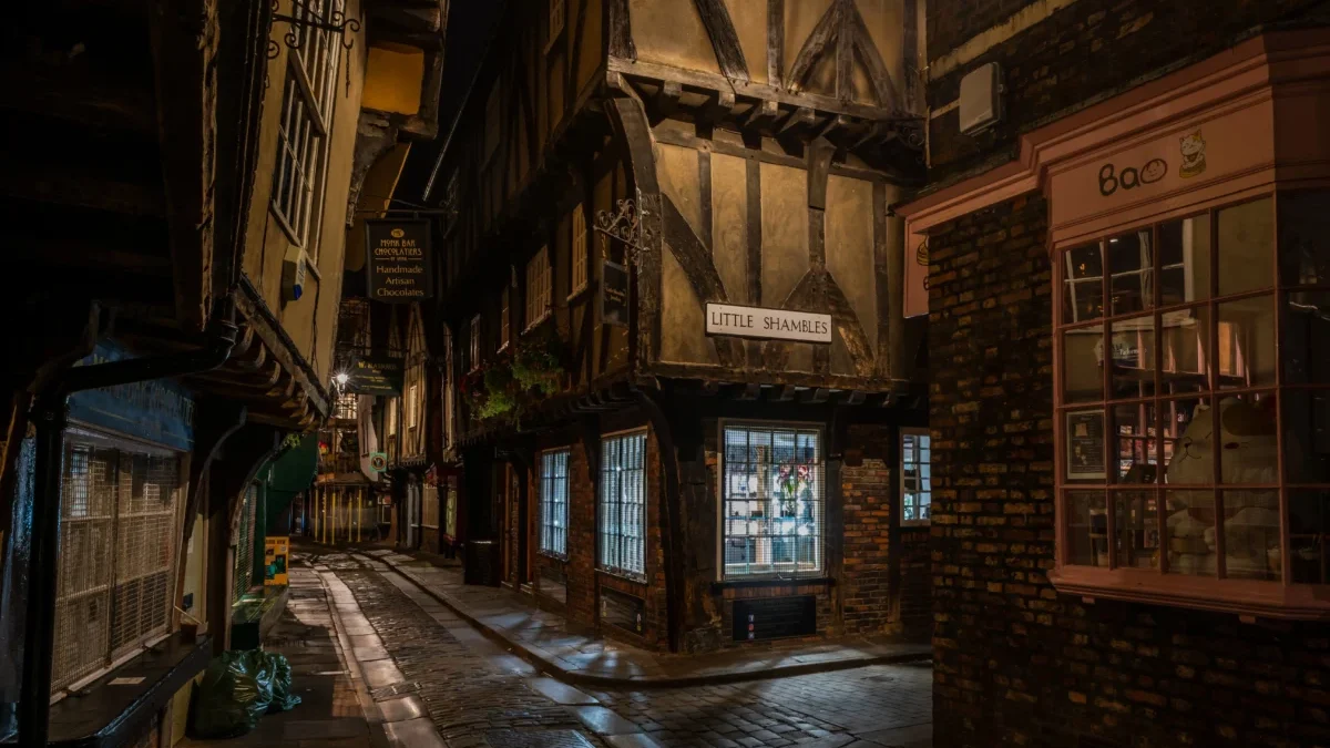 Narrow, cobblestone alley at night lined with timber-framed buildings and warmly lit storefronts, including a pink-brick shop on the right.