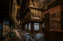 Narrow, cobblestone alley at night lined with timber-framed buildings and warmly lit storefronts, including a pink-brick shop on the right.