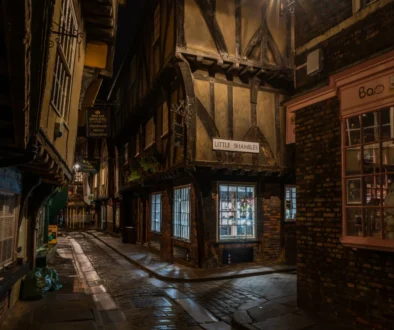 Narrow, cobblestone alley at night lined with timber-framed buildings and warmly lit storefronts, including a pink-brick shop on the right.